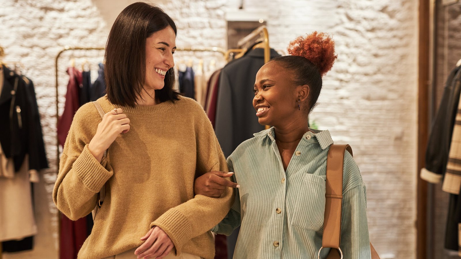 Deux amies bras dessus bras dessous dans une boutique de vêtements, l'une en pull moutarde, l'autre en chemise verte.