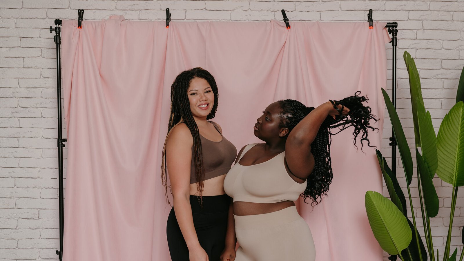 Deux femmes rondes en tenue de sport rient ensemble devant une toile de fond rose, dans un studio photo.