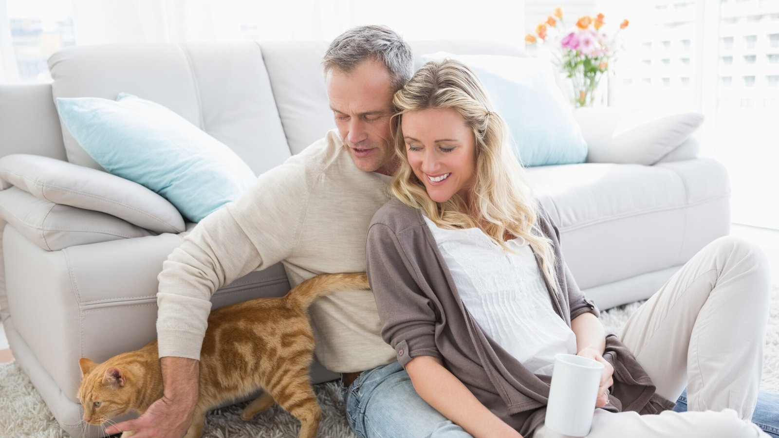 Un couple assis au pied d'un canapé caresse son chat roux, un café à la main.