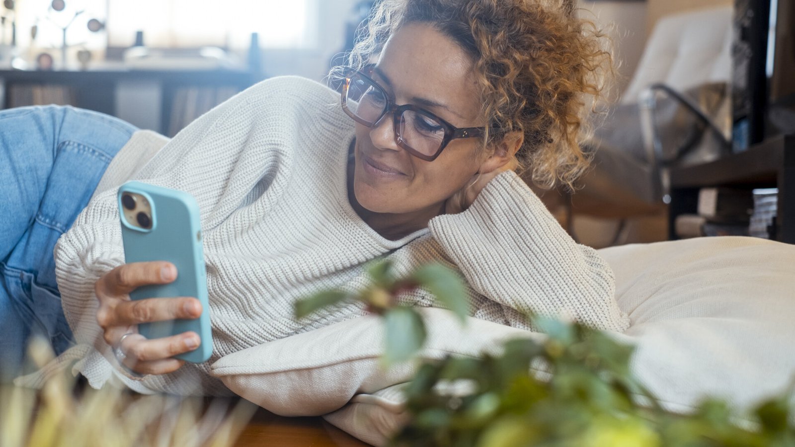 Une femme d'âge mûr aux cheveux bouclés et à lunettes consulte son téléphone, allongée sur un canapé.