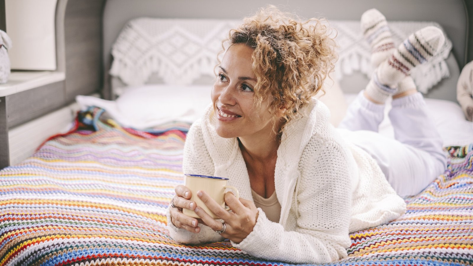 Une femme détendue, allongée sur un lit coloré, tient une tasse et sourit.