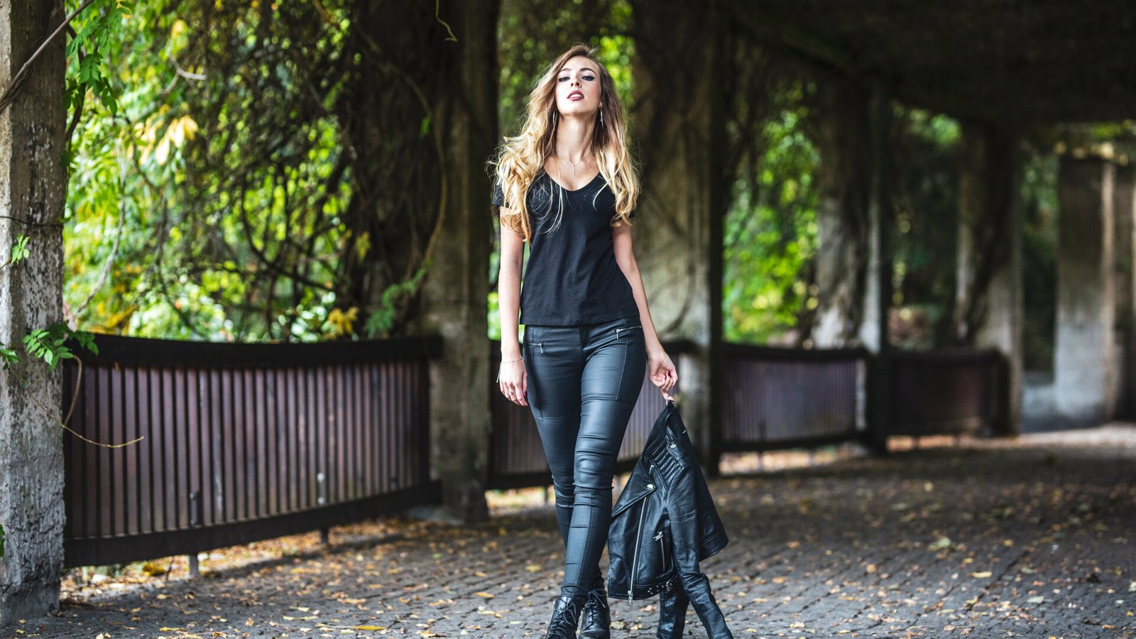 Une femme en pantalon de cuir noir et bottines marche sous une galerie couverte de végétation.