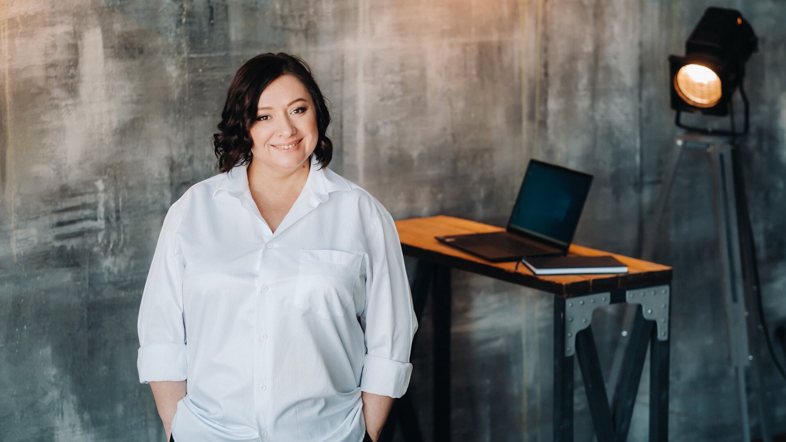 Une femme ronde en chemise blanche et pantalon noir, debout dans un bureau à l'ambiance loft.