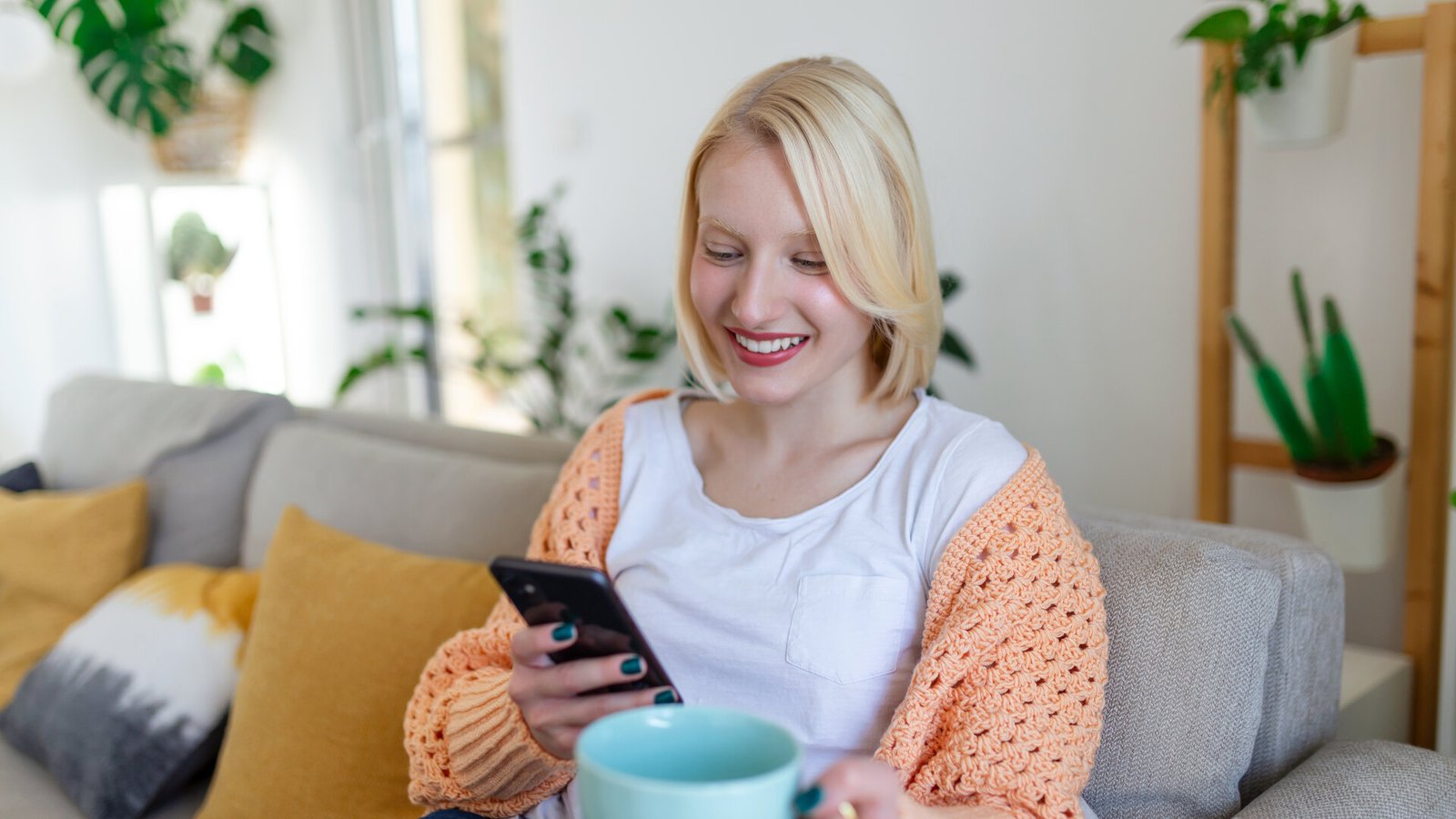 Une femme sourit devant son téléphone, installée sur un canapé, une tasse à la main.